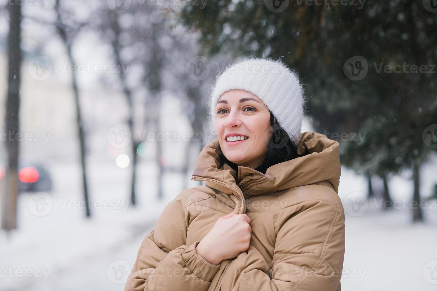 een gelukkig vrouw in een hoed is opwarming haarzelf in een jas. het staat in de stad in winter onder de sneeuw foto