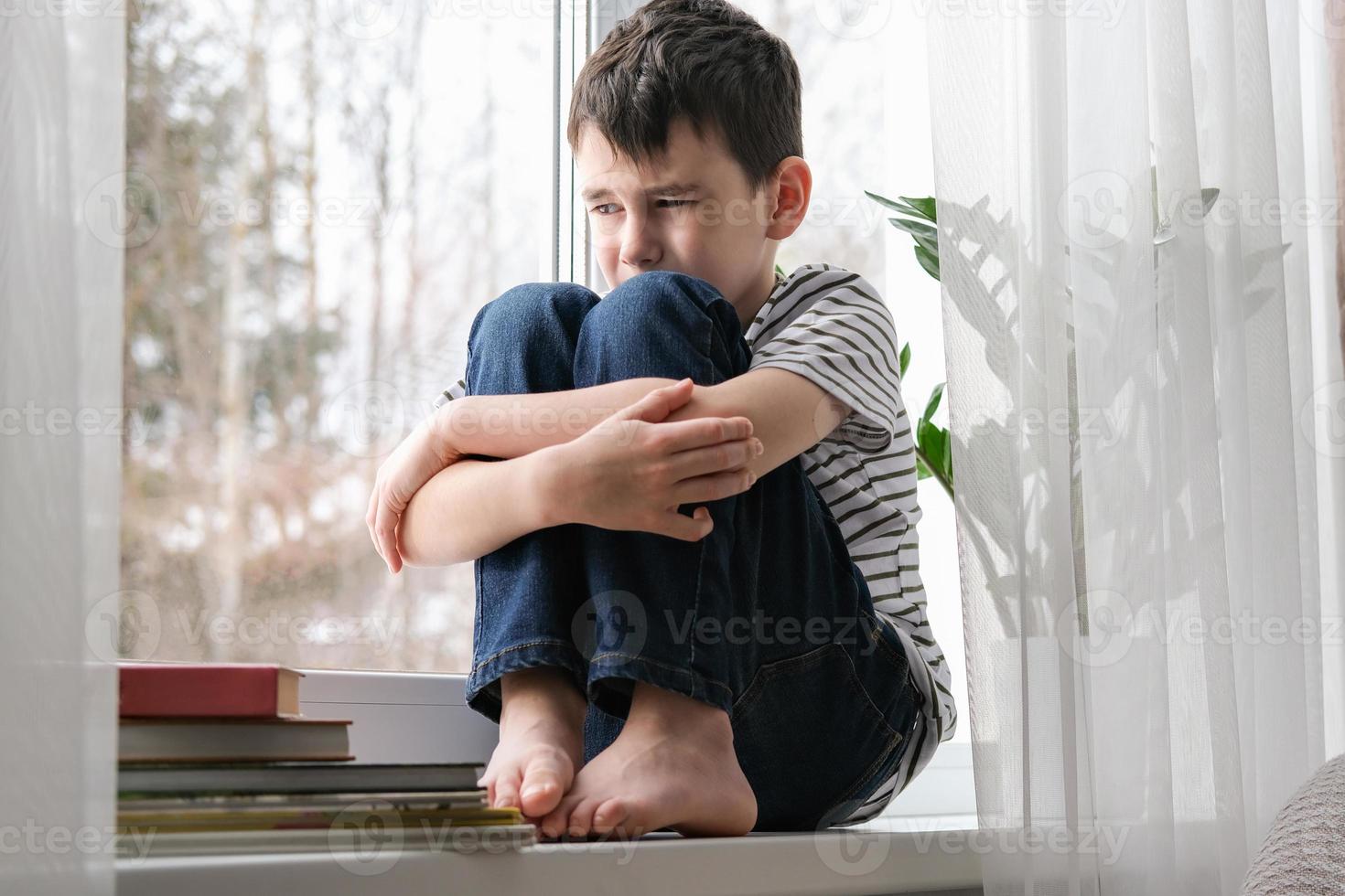 een verdrietig jongen zit Aan de vensterbank knuffelen zijn knieën. slecht stemming, depressie. de jongen is verdrietig alleen Bij huis foto