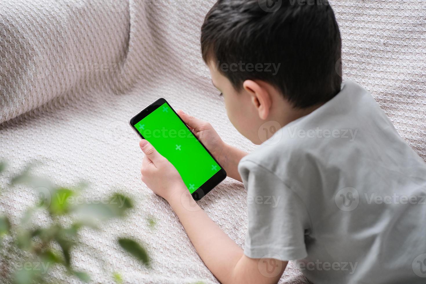 de jongen looks Bij de scherm van de telefoon met een chroom sleutel binnenshuis. een onherkenbaar kind toepassingen een smartphone met een groen scherm naar kijk maar een video. foto