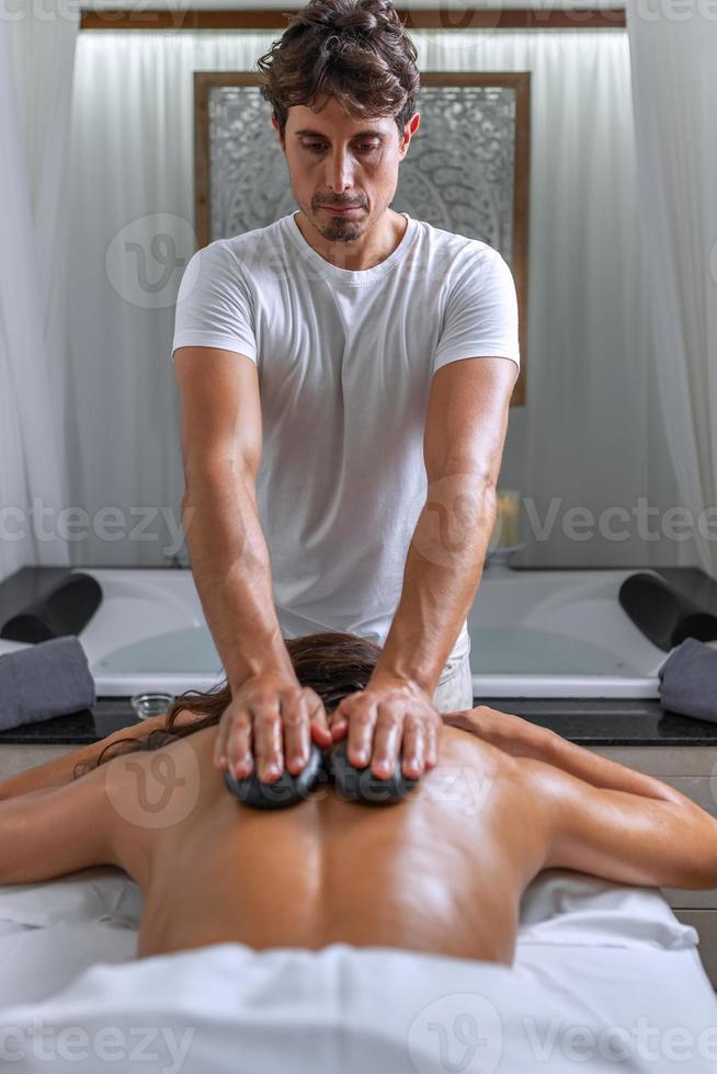 professioneel masseur het uitvoeren van een terug massage met stenen naar een vrouw in spa salon foto