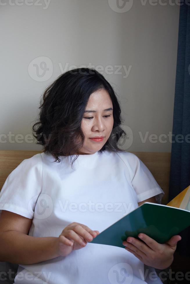 jong Aziatisch zwanger vrouw lezing boek Aan de bed Bij huis terwijl Holding haar buik foto