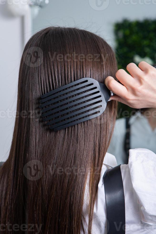 jong vrouw kammen haar lang donker haar- met een kam in een schoonheid salon. een Rechtdoor gezond brunette haar- dat heeft ondergaan de haar- rechttrekken procedure. foto