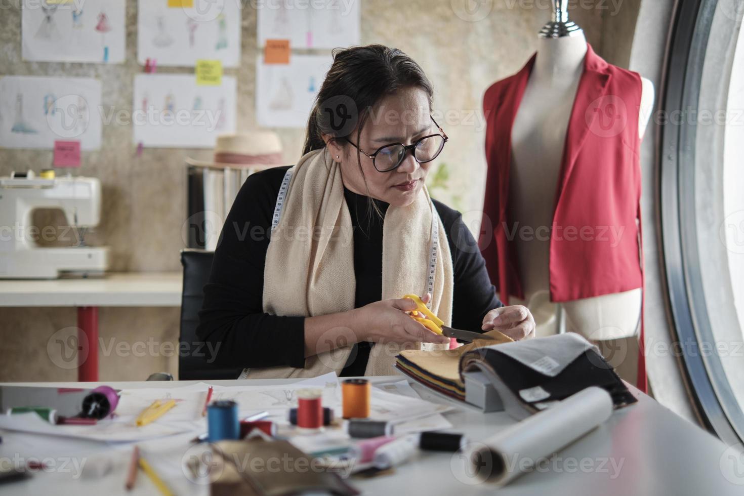 Aziatisch middelbare leeftijd vrouw mode ontwerper werken in studio, snijdend en kiezen kleding stof en draad kleuren met tekening schetsen voor jurk ontwerp collecties. professioneel winkel kleermaker ondernemer. foto