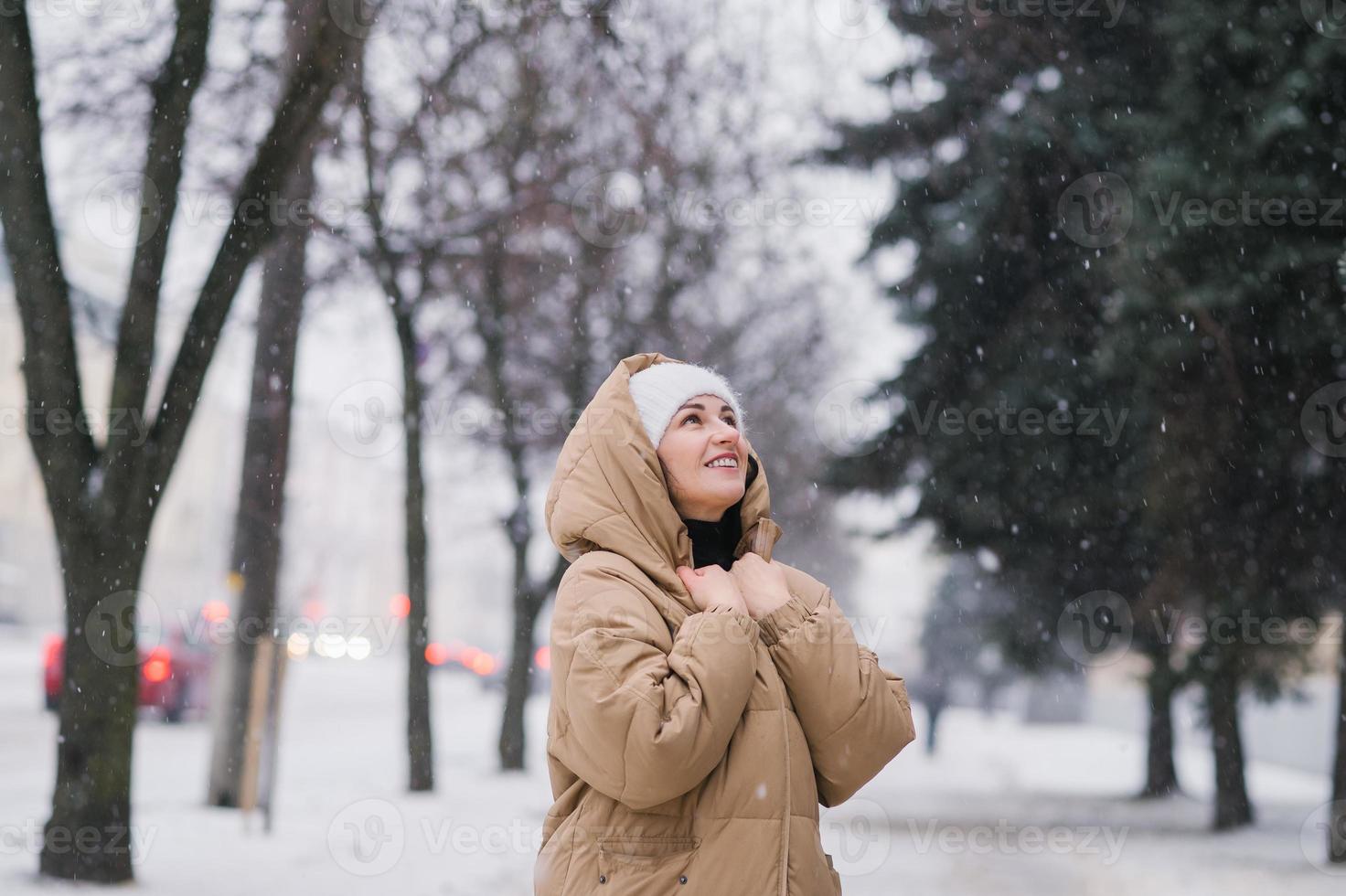 een gelukkig vrouw in een met een kap jas. het staat in de stad in winter onder de sneeuw foto