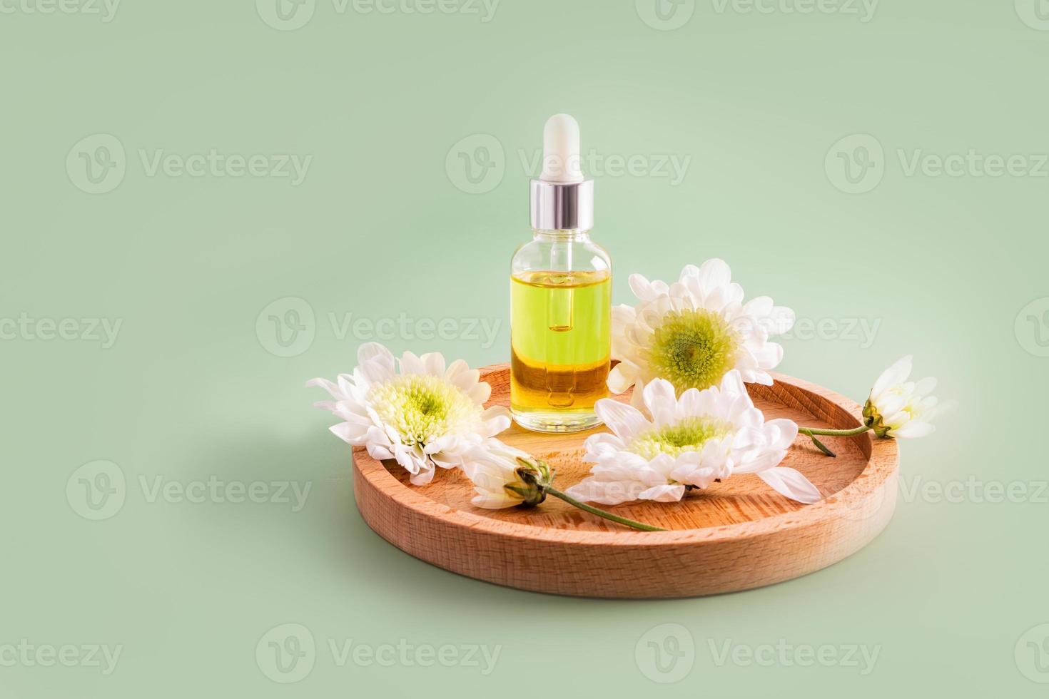 een glas fles met een pipet en een wit pet met een natuurlijk kunstmatig, serum of olie staat Aan een houten bord met bloemen. groen achtergrond. foto