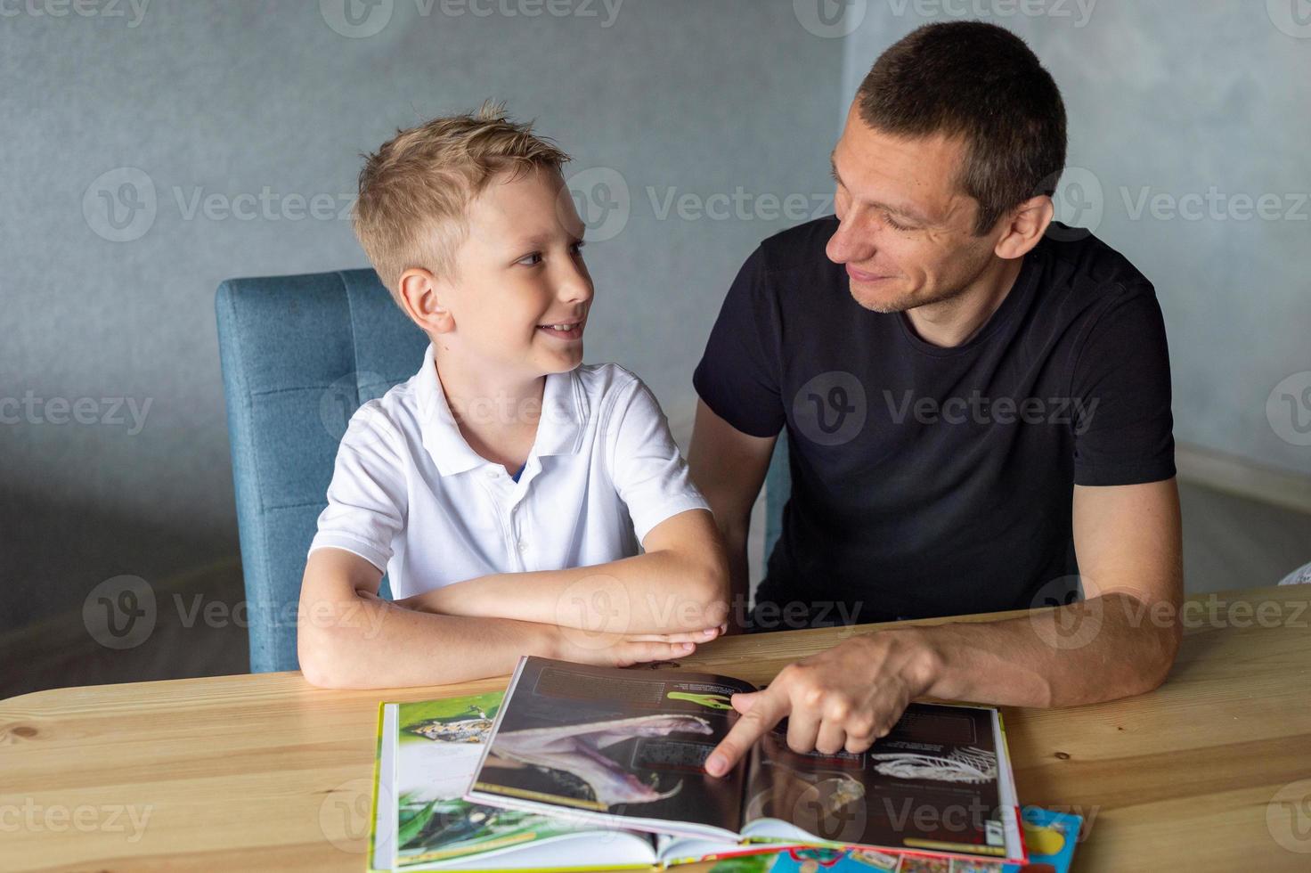 een schattig jongen is zittend Bij de tafel met zijn vader en aan het kijken een boek over slangen foto