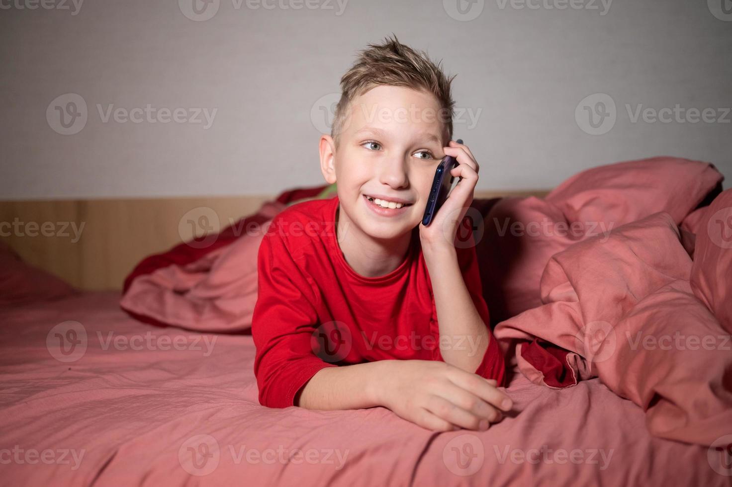 een schattig jongen in rood pyjama is aan het liegen in bed onder een deken en pratend Aan de telefoon foto