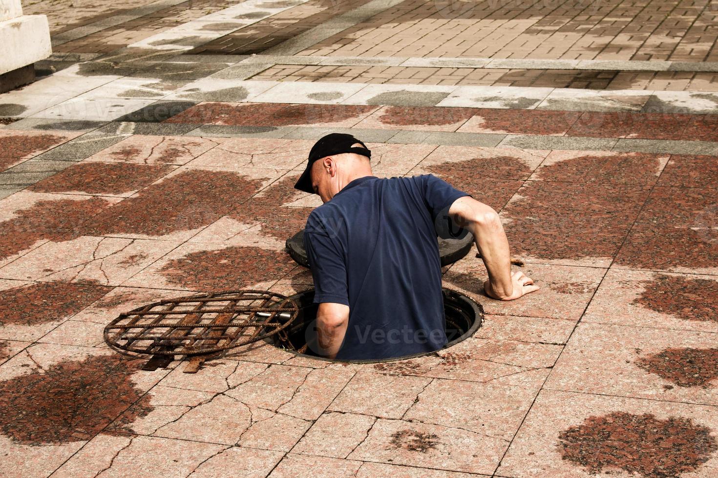 een arbeider klimt uit van een Open mangat in de weg. gevaarlijk Open onbeschermd mangat Aan de weg. ongeluk met een riool mangat in de stad. de concept van repareren ondergronds Gereedschap. foto