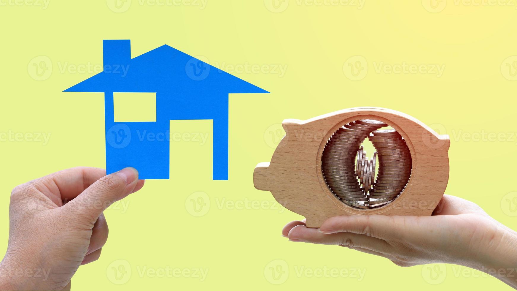 handen met een modelhuis en een houten spaarvarken met muntstapels die binnen op gele achtergrond worden geplaatst foto