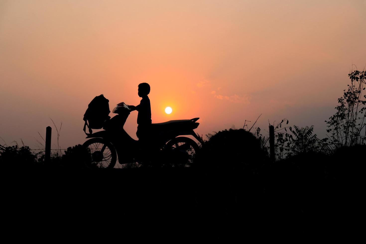 silhouet van kind op bromfiets met laagstaande zon aan de hemel foto