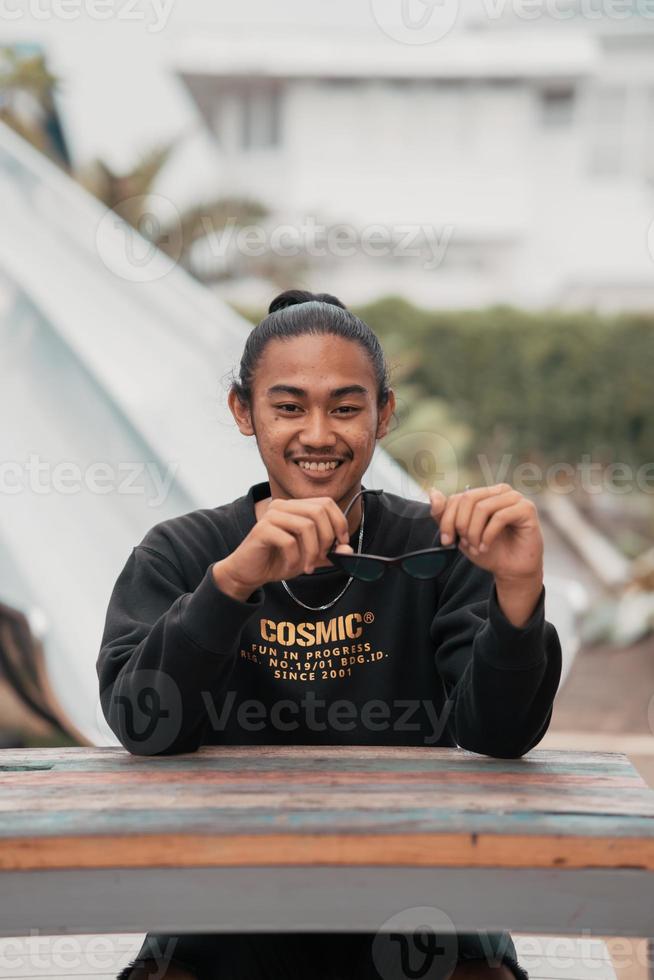 een Aziatisch Mens met zijn haar- gebonden omhoog en vervelend een zwart trui jasje is zittend ontspannende Bij een cafe tafel wanneer vergadering zijn vrienden foto