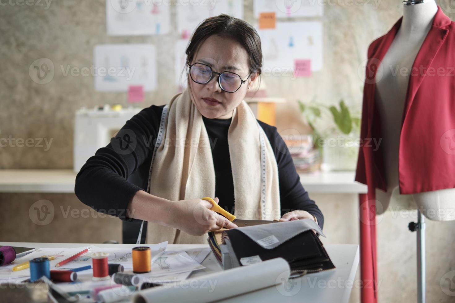 Aziatisch middelbare leeftijd vrouw mode ontwerper werken in studio, snijdend en kiezen kleding stof en draad kleuren met tekening schetsen voor jurk ontwerp collecties. professioneel winkel kleermaker ondernemer. foto