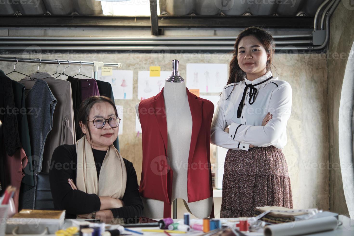 mode team mensen, Aziatisch vrouw ontwerper en tiener assistent in studio, armen gekruiste en glimlach, gelukkig werken met draad en naaien voor jurk ontwerp, professioneel winkel kleermaker mkb ondernemer. foto