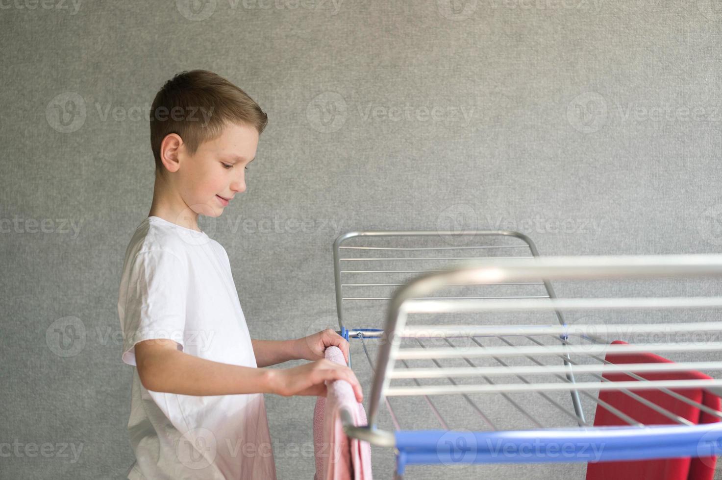 schattig jongen hangende een nat handdoek Aan een metaal kleren droger, huiswerk, huishouden klusjes foto