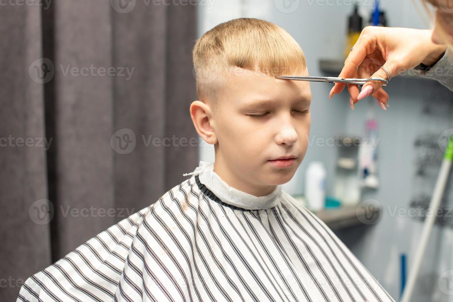 een schooljongen is zittend in een kapperszaak, aan het doen zijn haar- met schaar voor kapsels foto