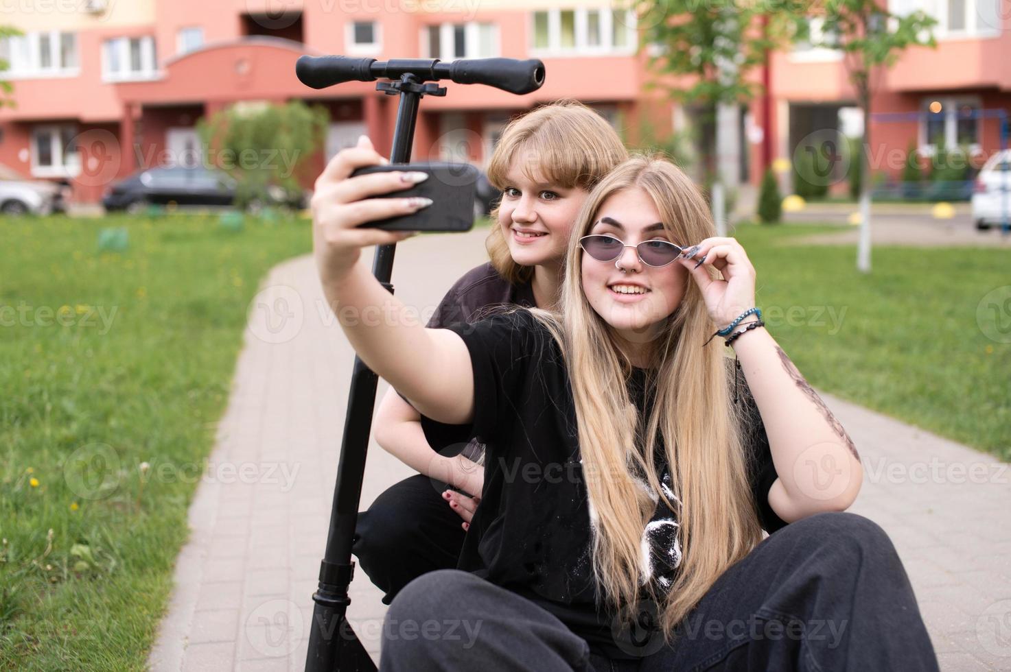 twee grappig meisjes zijn zittend Aan een scooter in de stad en nemen selfies Aan hun telefoon. tieners glimlach foto