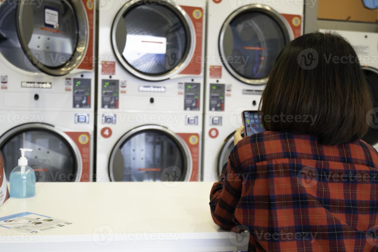 achterzijde visie van vrouw zittend met mobiel telefoon aan het wachten voor wasserij gewassen in automatisch het wassen machine Bij wasserette winkel. rij van industrieel het wassen machine in de munt wasserij openbaar wasserette. foto