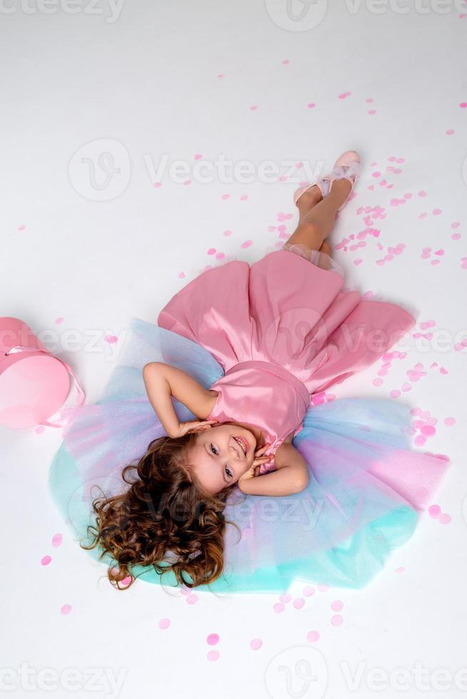 mooi weinig meisje in een chique roze jurk leugens Aan de verdieping bezaaid met confetti. top visie. mode en stijl. kind viert zijn verjaardag. foto in de studio. ruimte voor tekst. hoog kwaliteit foto
