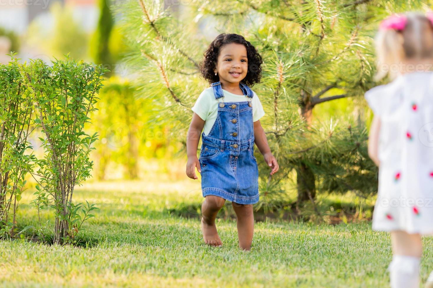 portret weinig gelukkig speels kleuter donker meisje in een denim zonnejurk staand in de tuin Aan een zonnig dag. wandelen in de vers lucht. concept van een gelukkig jeugd. ruimte voor tekst. hoog kwaliteit foto