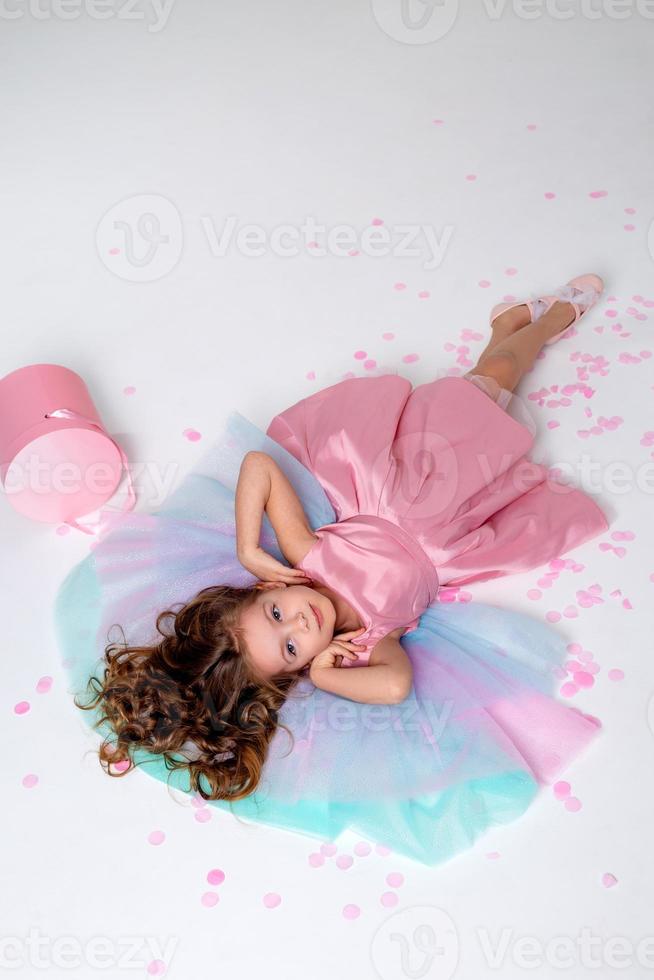 mooi weinig meisje in een chique roze jurk leugens Aan de verdieping bezaaid met confetti. top visie. mode en stijl. kind viert zijn verjaardag. foto in de studio. ruimte voor tekst. hoog kwaliteit foto