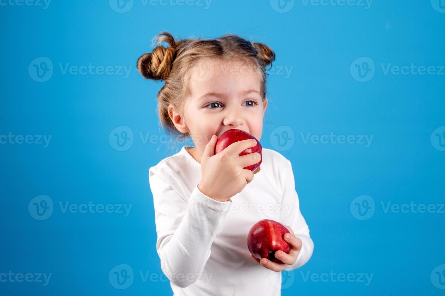 schattig weinig meisje met krullen in een gestreept jurk is Holding een groot rood appel in haar handen. blauw achtergrond. nuttig producten voor kinderen. gezond tussendoortje. ruimte voor tekst. hoog kwaliteit foto