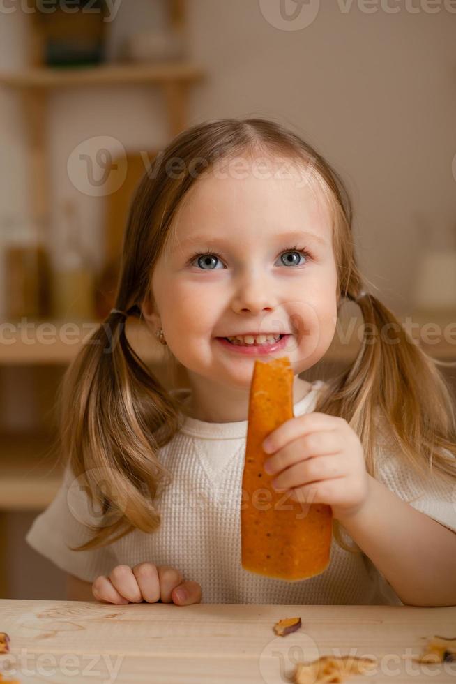 schattig weinig meisje eet natuurlijk pastille Bij huis in een houten keuken. voedsel voor kinderen van natuurlijk producten foto