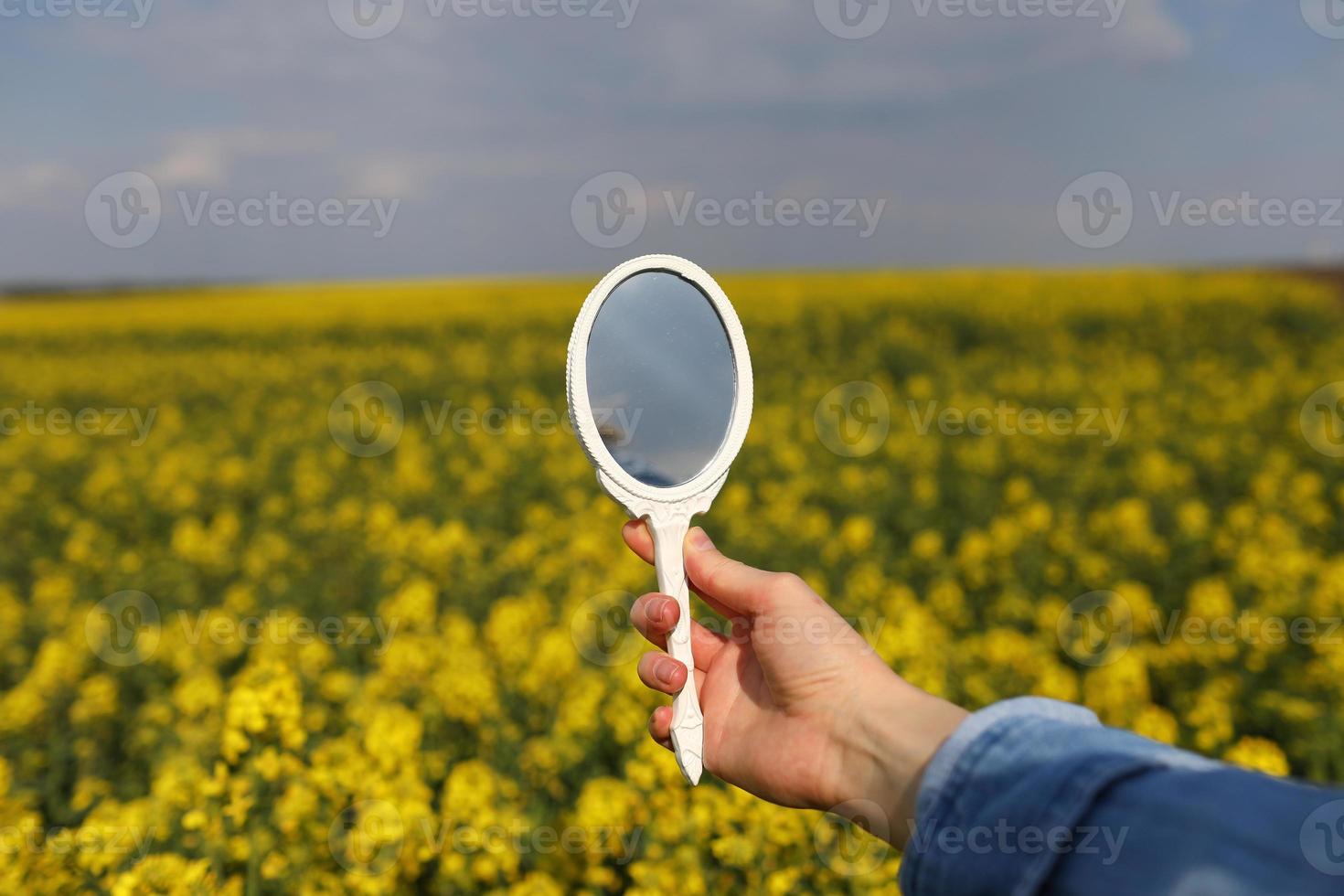 een vrouwelijke hand met een miniatuur retro spiegel op het gebied van gele wilde bloemen wordt weerspiegeld. creatief zomer helder concept. foto