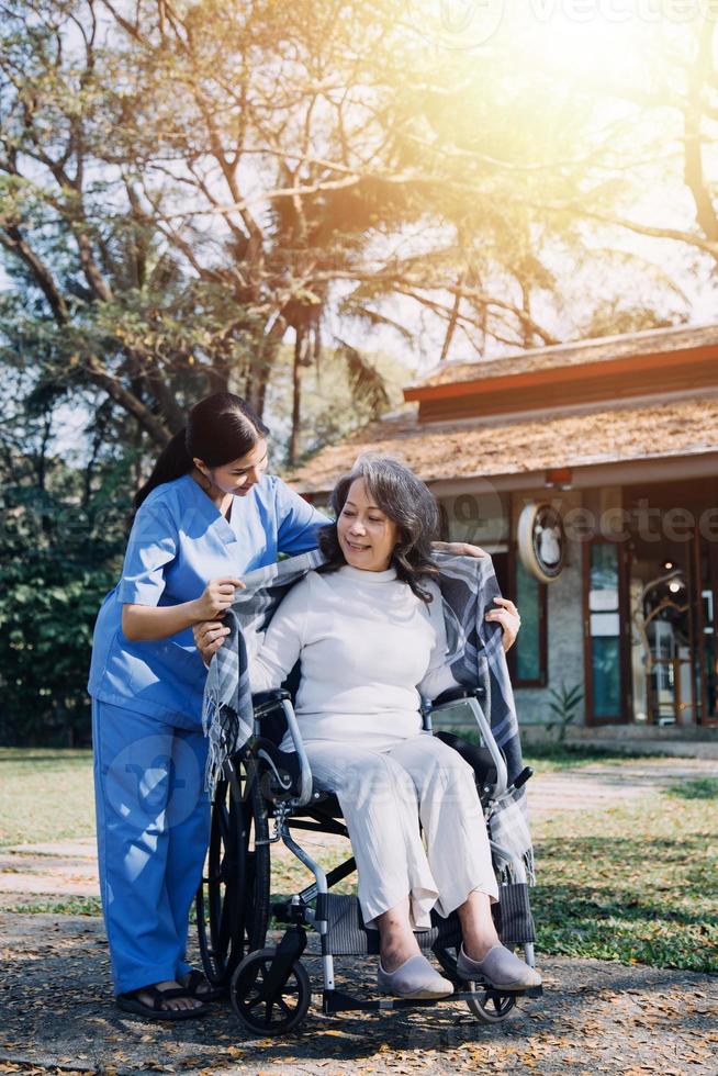 Aziatisch jong verzorger verpleegster ondersteuning senior ouder mannetje wandelen buitenshuis. specialist meisje dokter helpen en nemen zorg van ouderen volwassen Mens geduldig aan het doen fysiek behandeling in openbaar park Bij verpleging huis. foto