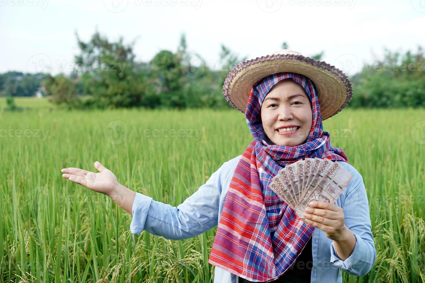 Aziatisch boer vrouw is Bij rijstveld veld, draagt hoed en rood plaid shirt, houden Thais bankbiljet geld. concept , boer gelukkig naar krijgen winst, inkomen, landbouw ondersteunen geld. foto