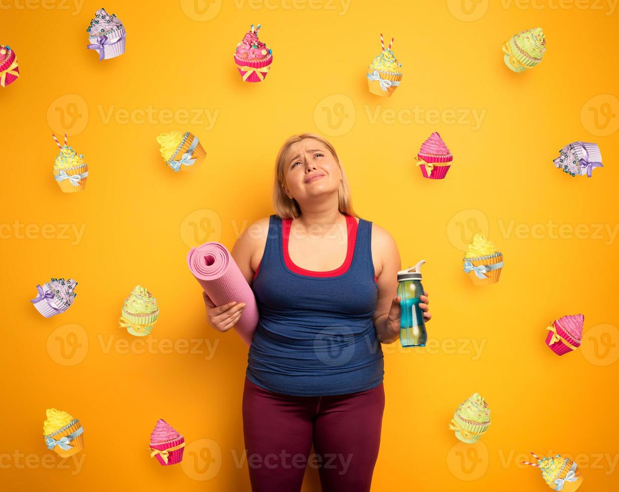 dik meisje denkt naar eten snoepgoed in plaats daarvan van Doen Sportschool. geel achtergrond foto