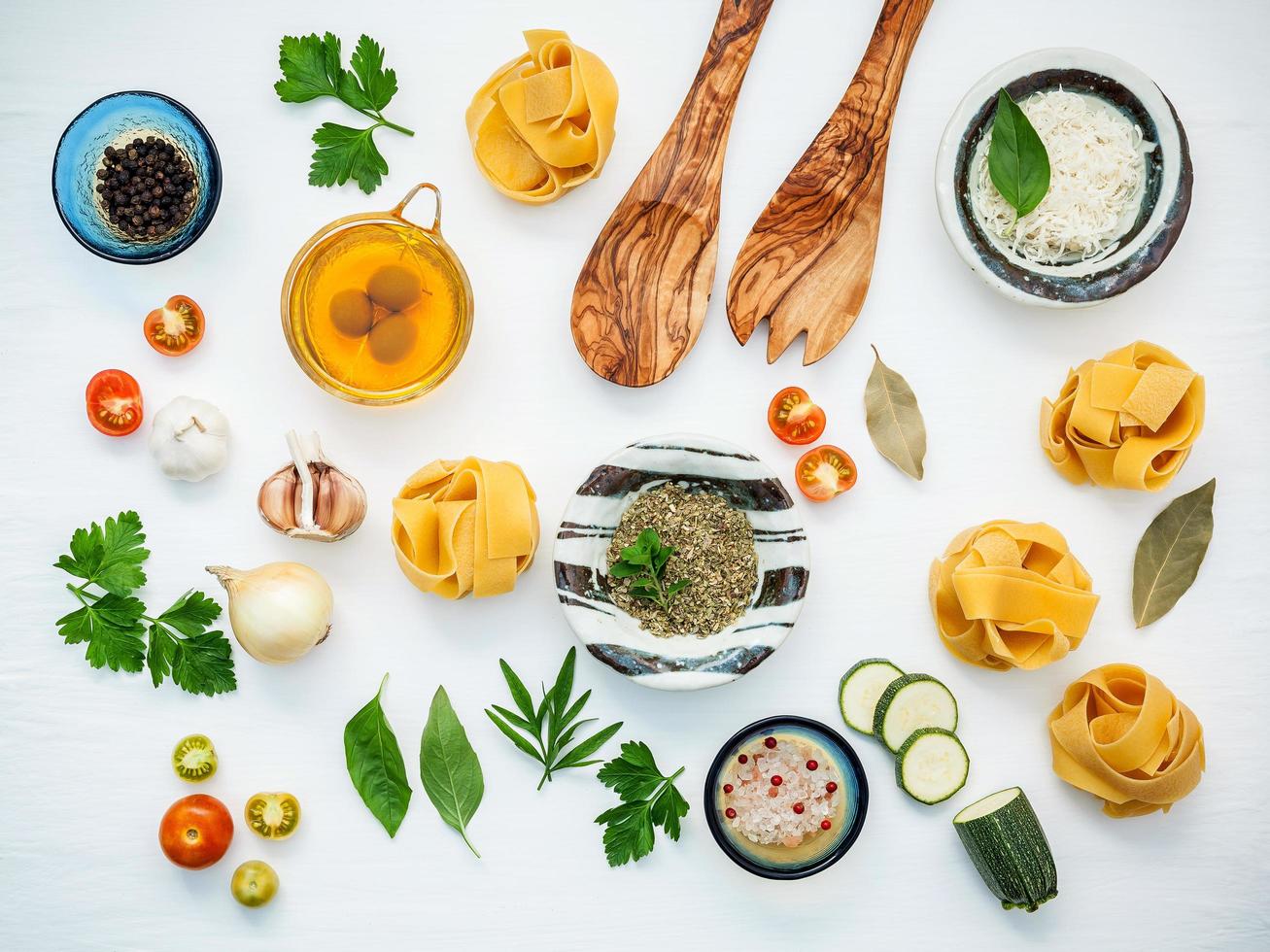 Italiaans voedselconcept en menuontwerp. gedroogde fettuccine met houten spatel en ingrediënten zoete basilicum, tomaat, knoflook, peterselie, laurierblaadjes, peper en courgette op witte houten vlakke achtergrond. foto