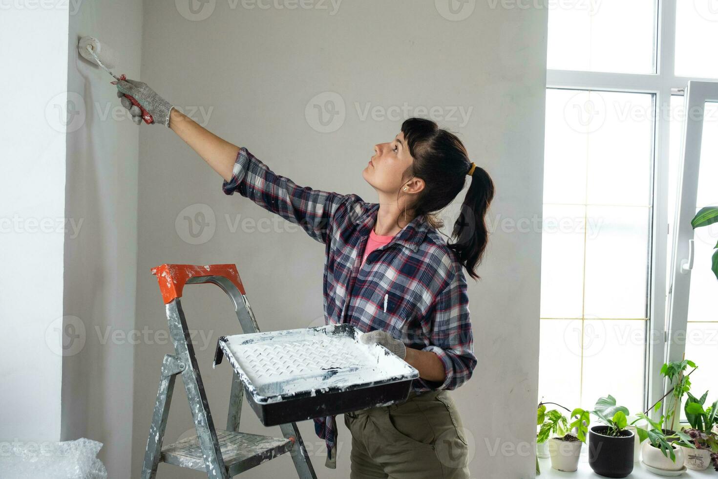 vrouw in verf rol en wit verf verven de muur van huis Aan trapladder. bouw werk en kunstmatig reparaties in huis, muur schilderen, verven, afwerking werk met uw eigen handen foto