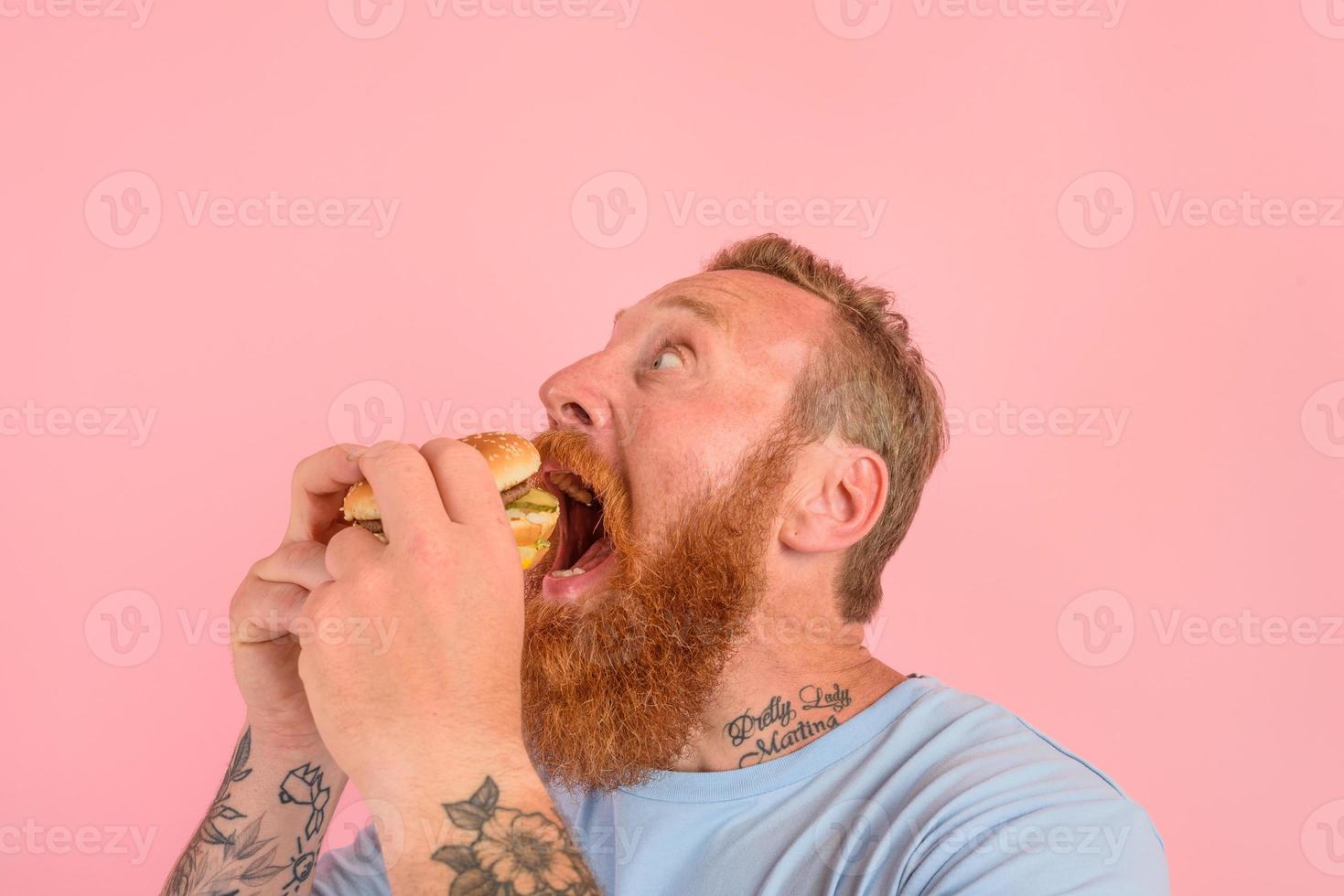 hongerig Mens met baard en tatoeages eet een sandwitch met Hamburger foto