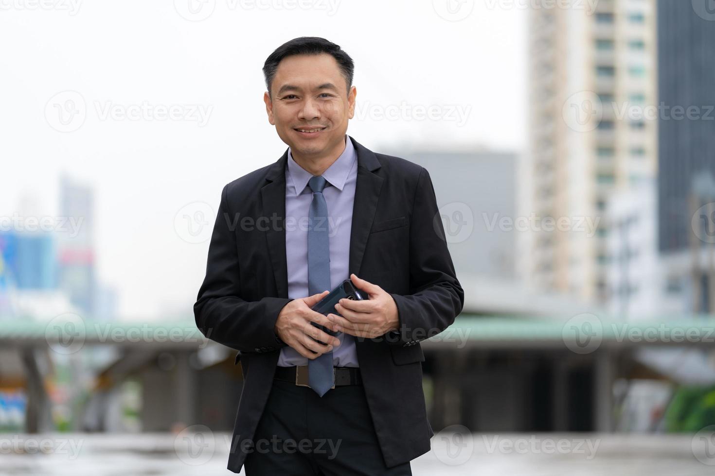 Aziatische zakenman permanent en bedrijf mobiele telefoon met zakelijke kantoorgebouwen op de achtergrond van de stad foto