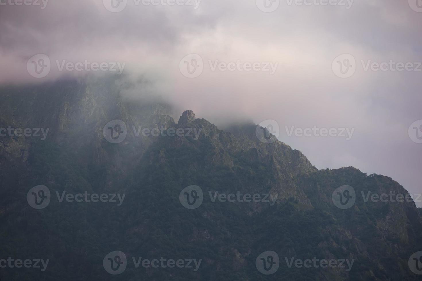 donker sfeervol surrealistische landschap met een donker rotsachtig berg top in laag wolken in een grijs bewolkt lucht. foto