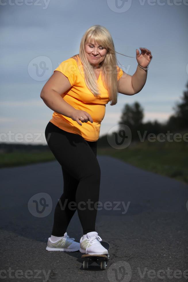 mollig middelbare leeftijd Europese vrouw, overgewicht, in een trainingspak, leert naar rijden een skateboard, gaat in voor sport. foto