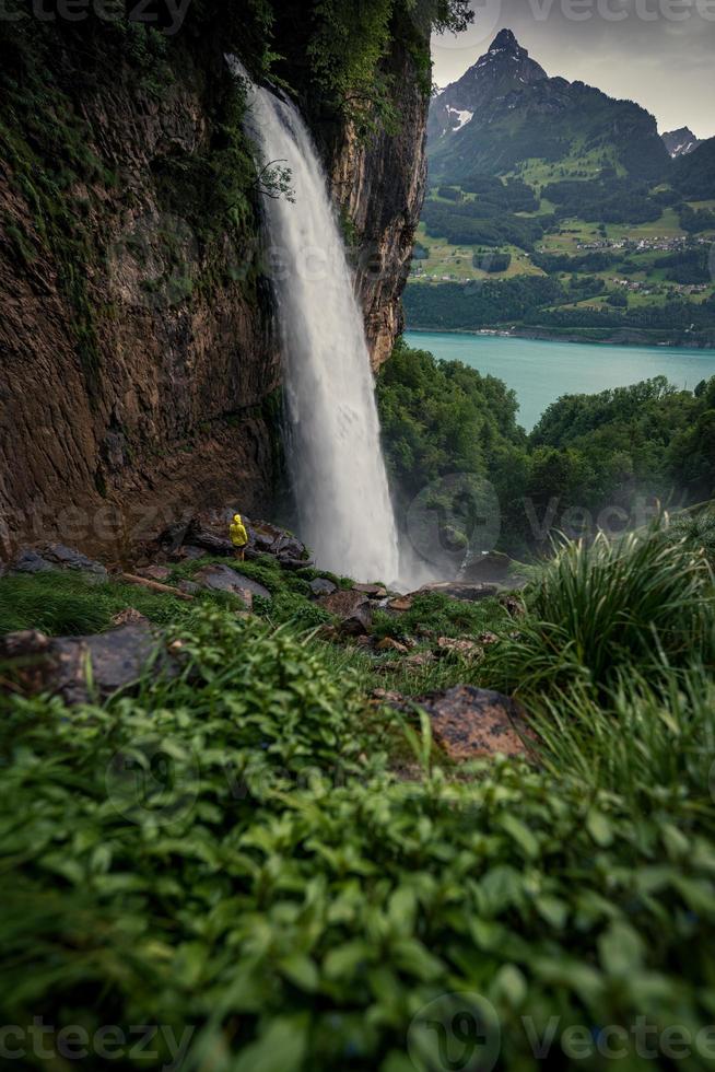 een machtig waterval vloeiende uit van een berg omringd door groen gras, een persoon in een geel regenjas staand in voorkant van het foto