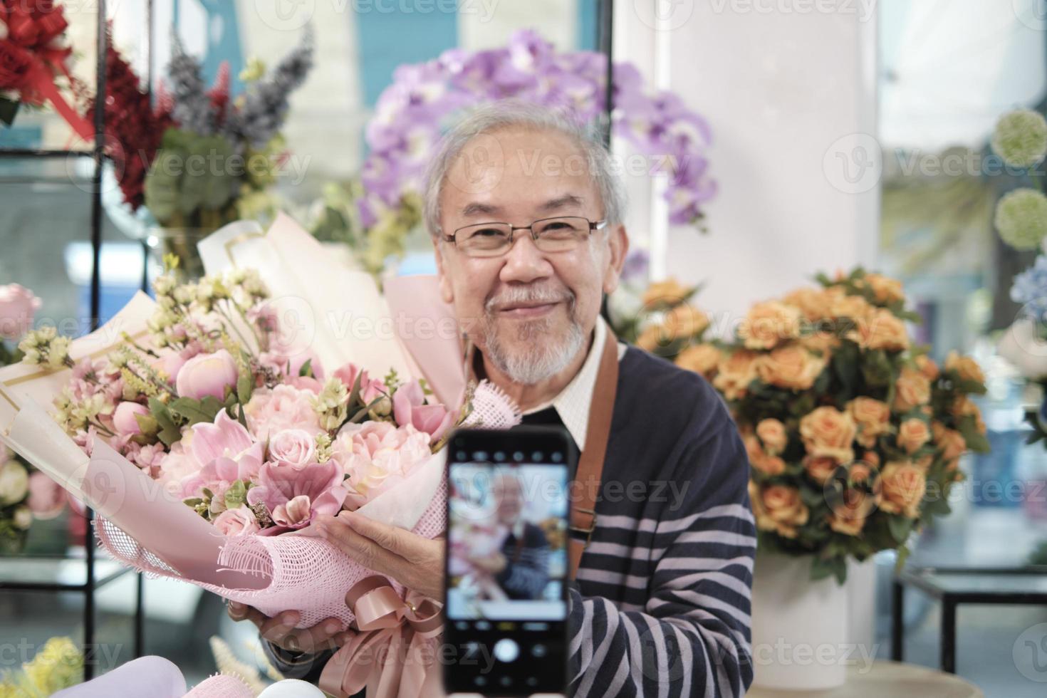 e-commerce bedrijf, een senior mannetje bloemist arbeiders demonstreren en tonen bloemen regelingen via online leven streaming met smartphone toepassing in een helder bloem winkel, een mooi bloesems op te slaan. foto