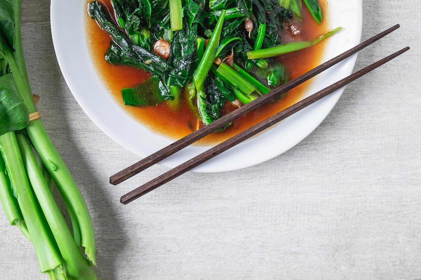 gebakken chinese boerenkool met oestersaus met stokjes naast verse chinese boerenkool op een witte plaat op houten tafel foto