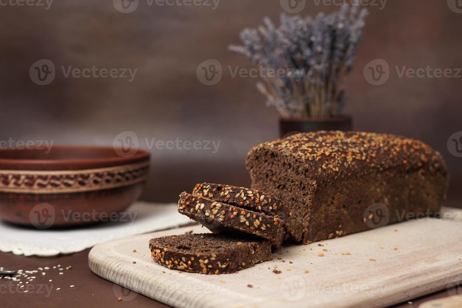 zwart brood met granen gesneden Aan een houten snijdend bord tegen een bruin achtergrond. De volgende naar de brood is een klei bord, vork, lepel en wit katoen tafelkleed foto