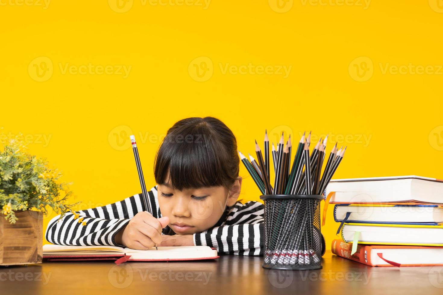 jong Aziatisch meisje dat in een notitieboekje schrijft naast stapel boeken, kop potloden en bloemen met gele achtergrond foto