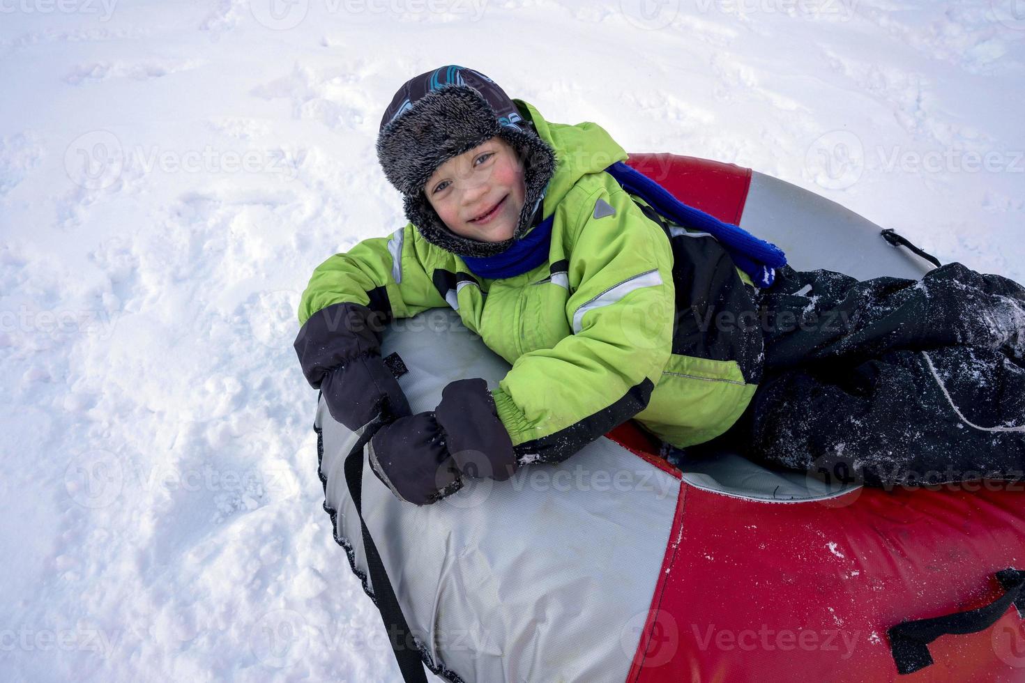 grappig Kaukasisch jongen 5 jaren oud ritten in winter Aan een groot opblaasbaar kwarktaart foto