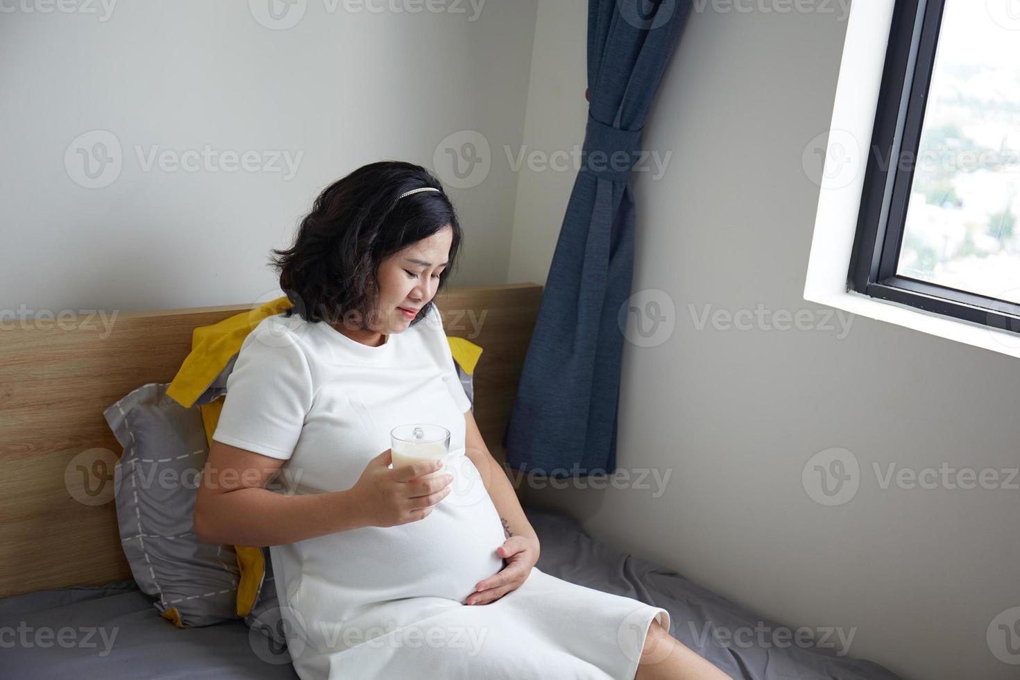 gelukkig jong Aziatisch zwanger vrouw drinken melk Aan bed Bij huis terwijl Holding haar buik en denken foto