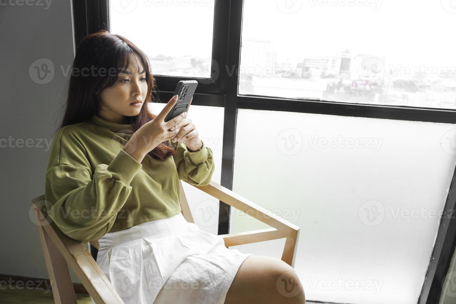 een jong Aziatisch vrouw is gevoel echt en gefocust Aan Holding haar smartphone in hand- terwijl zittend in een slaapkamer. foto