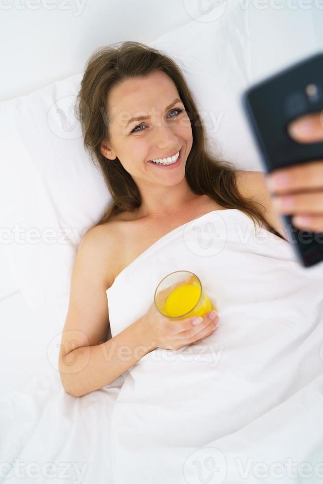 schattig vrouw cheques de smartphone en drankjes oranje sap in bed in de ochtend- foto