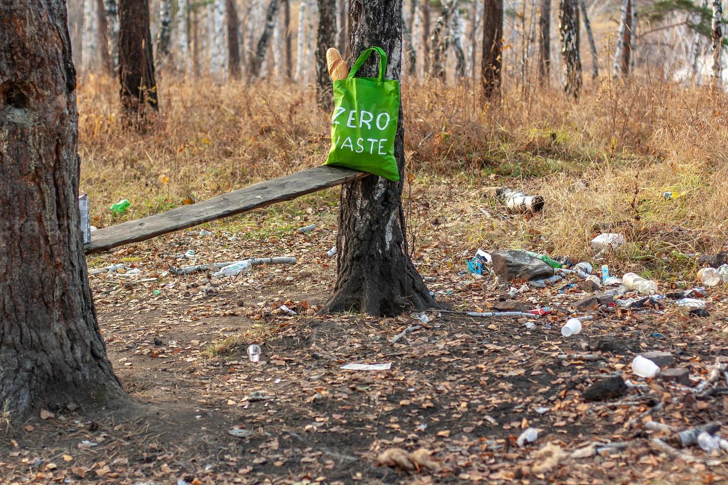 een textiel groen zak met de opschrift nul verspilling staat in de Woud De volgende naar de vuilnis verspreide Aan de grond. een veel van plastic gerechten en flessen van alcohol. selectief focus. foto