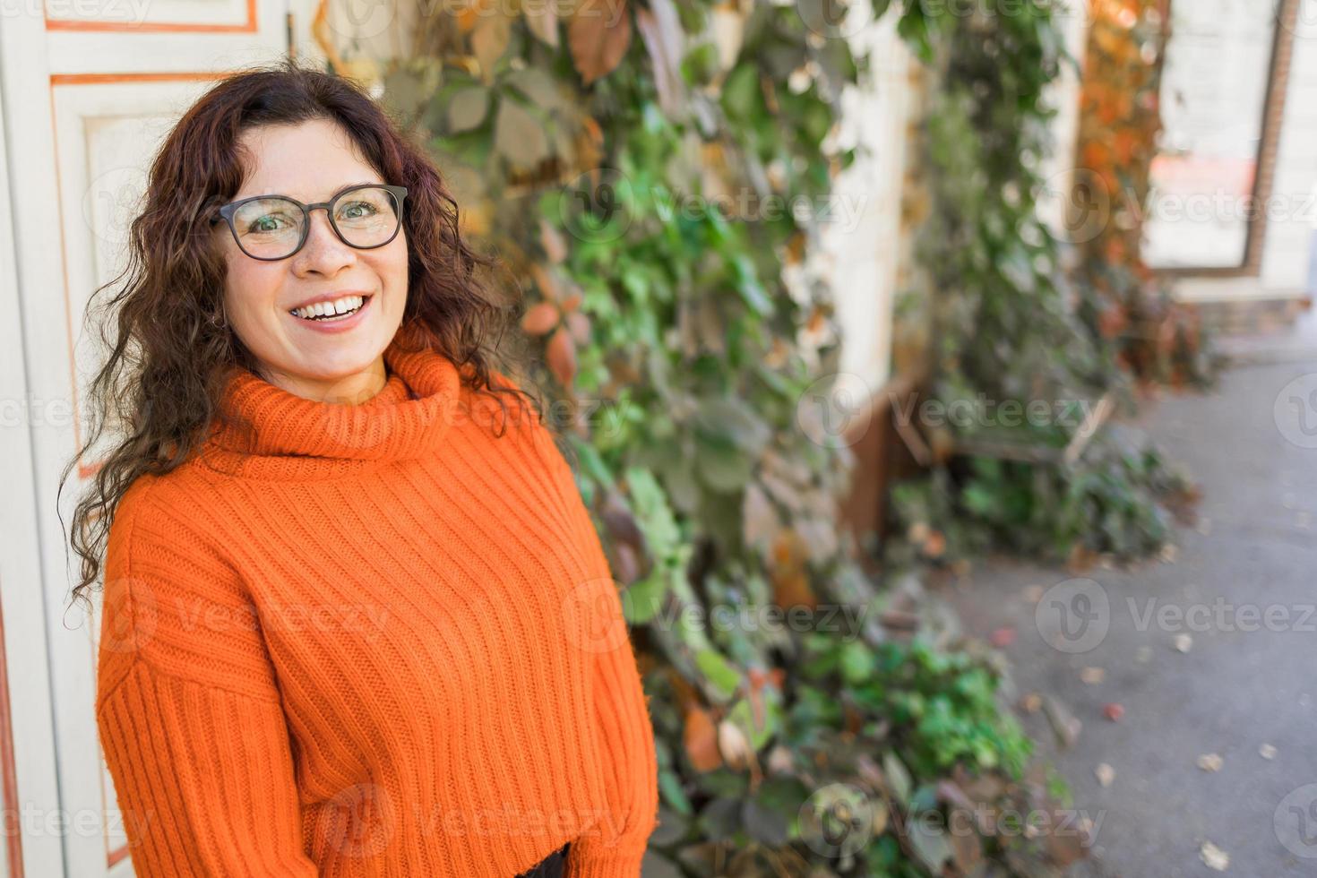 elegant gelukkig jong vrouw vervelend oranje sweater en bril. portret van glimlachen meisje in modieus bril in stad, kopiëren ruimte en leeg ruimte voor reclame foto