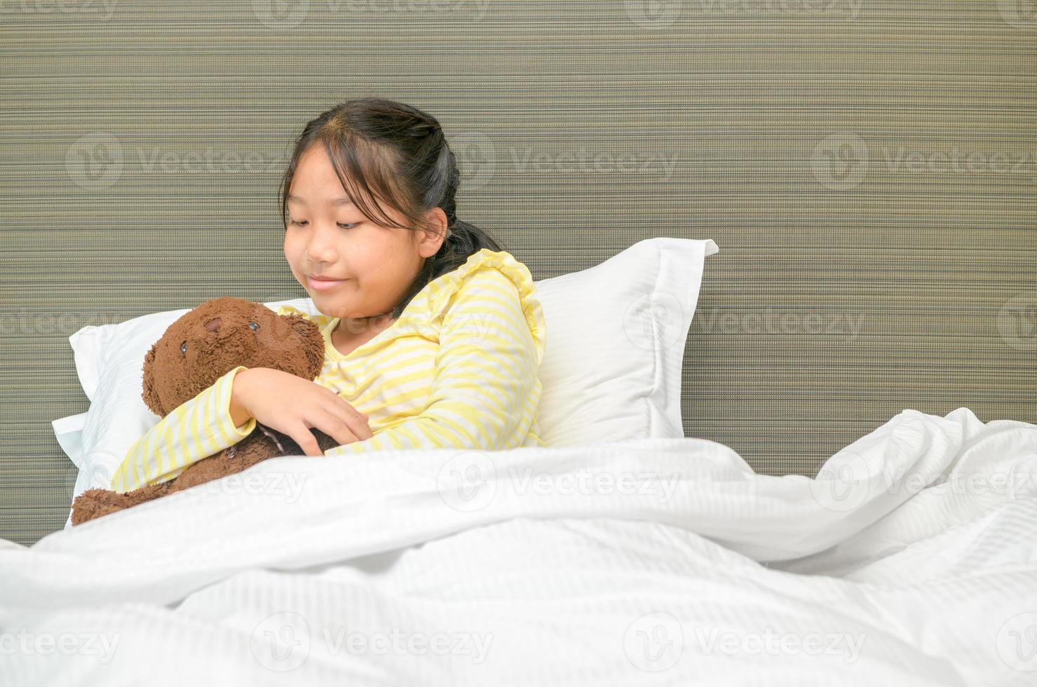 zoet weinig meisje is knuffelen een teddy beer, op zoek Bij beer en glimlachen terwijl aan het liegen Aan haar bed foto