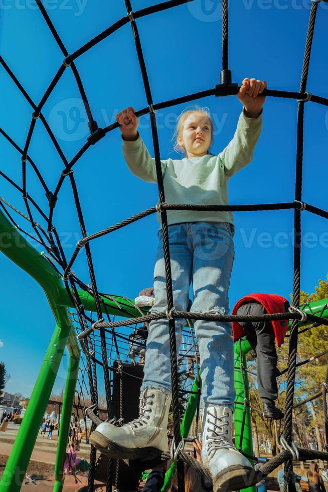 een gelukkig actief tien jaar oud meisje beklommen een touw web Aan een speelplaats tegen een helder blauw lucht Aan een zonnig dag foto