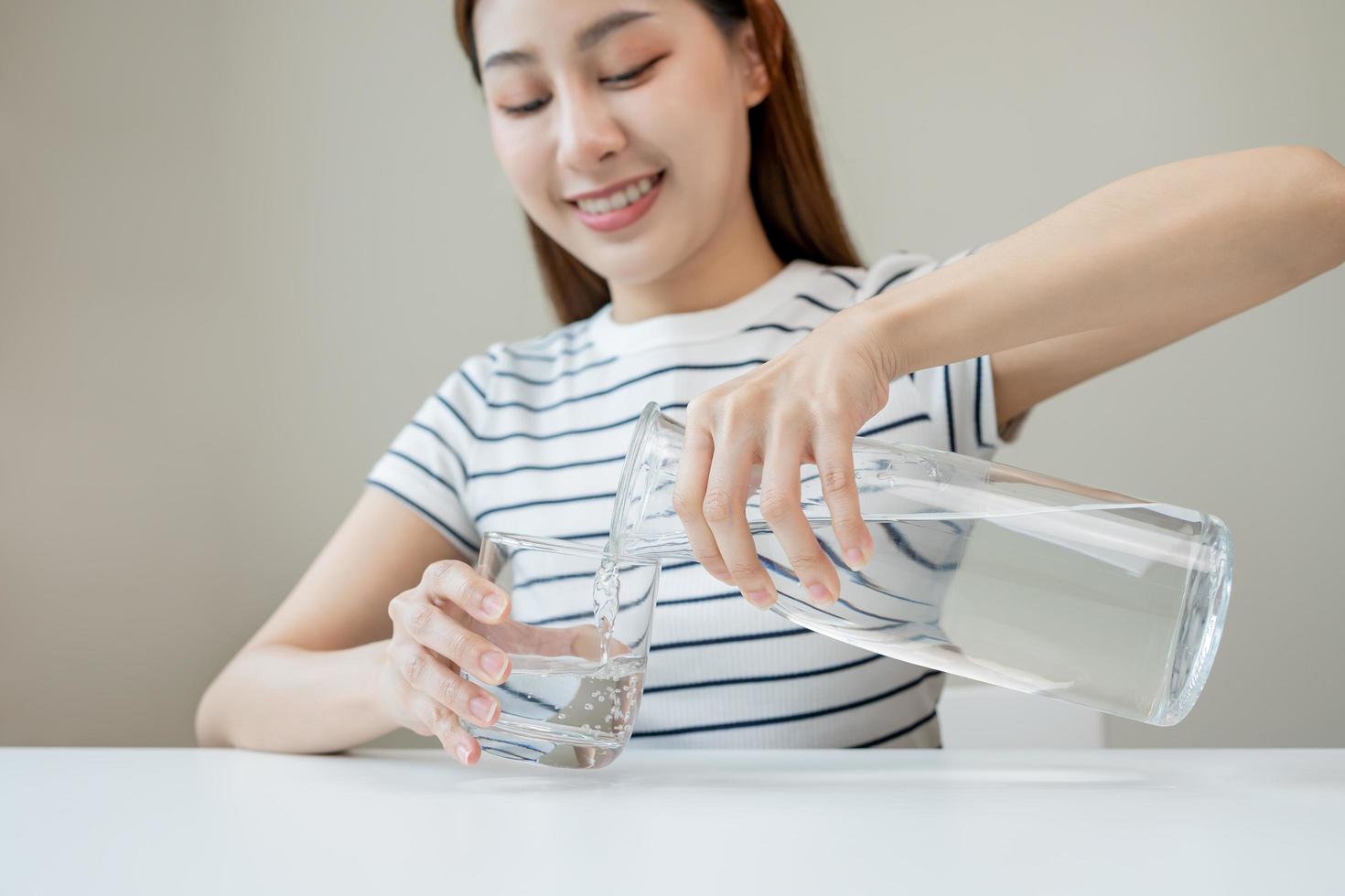 gelukkig mooi, dorstig Aziatisch jong vrouw, meisje drinken, hand- in vasthouden, gieten water van kruik in transparant glas van werper voor hydratatie van lichaam. Gezondheid zorg, gezond levensstijl concept. foto